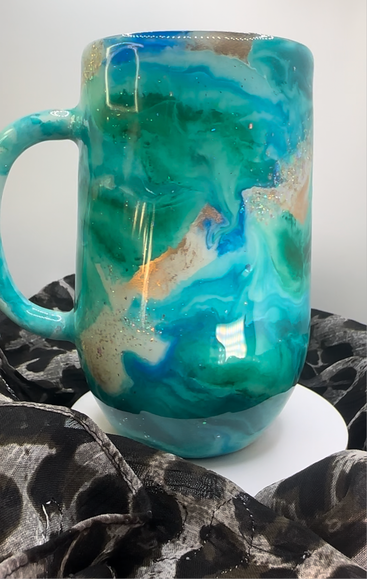 Turquoise tumbler with swirling blue, white, and gold accents, featuring a glossy finish, displayed on a white stand with black fabric.