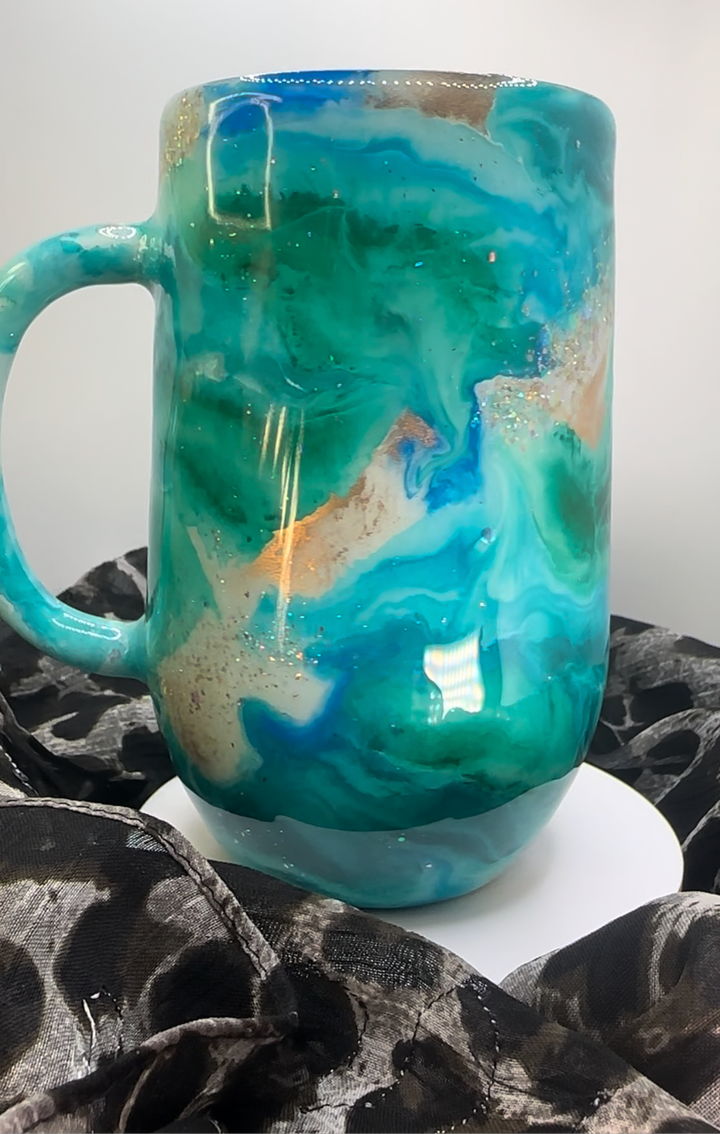 Turquoise tumbler with swirling blue, white, and gold accents, featuring a glossy finish, displayed on a white stand with black fabric.