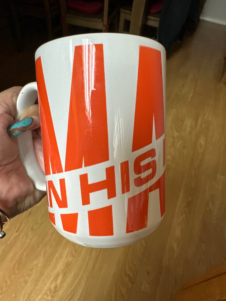 White ceramic mug with bold red block text partially visible, held by a hand with turquoise nails on a wooden floor background.