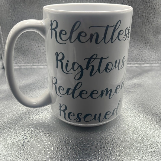 White ceramic mug with black script text reading "Relentless, Righteous, Redeemed, Rescued," placed on a metallic textured surface.