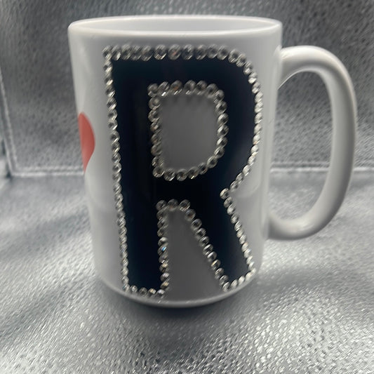 White ceramic mug featuring a bold black "R" outlined with sparkling rhinestones, set against a metallic textured background.